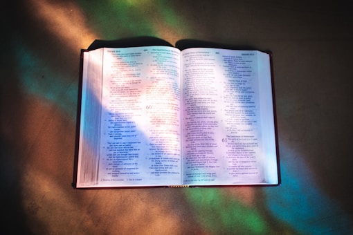 An open Bible glowing in the reflection of stained glass windows in a church. 