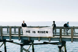 People Holding a He Is Risen Banner over the Side of a Boardwalk  image 1