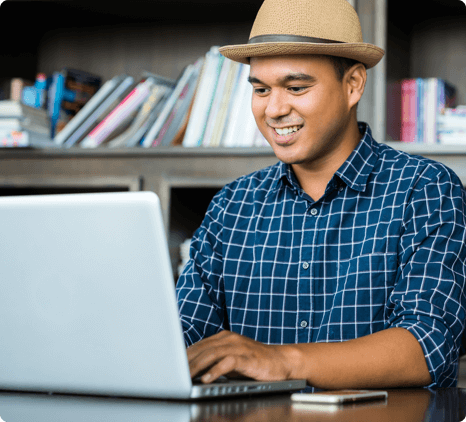 A man who appears to be in his 30s is sitting at a table with a bookshelf behind him, perhaps at a home office. He's cheerfully working on his laptop. There's an iPhone next to the laptop, face down on the table.