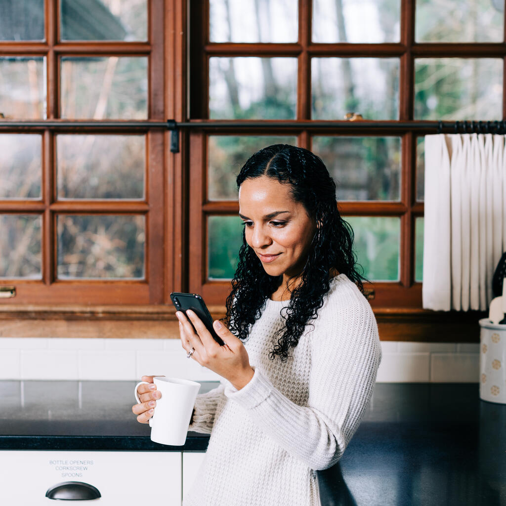 Woman Looking at Her Phone with a Cup of Coffee in the Kitchen large preview