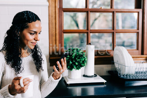 Woman Looking at Her Phone with a Cup of Coffee in the Kitchen