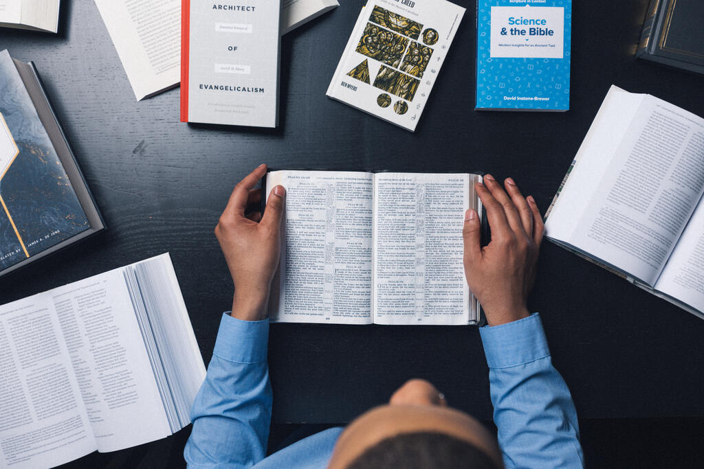 Seminary Student Reading the Bible Surrounded by Other Books and Commentaries large preview