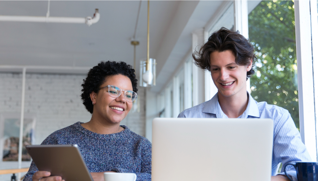 Man and woman sitting at a table together having a discussion and both looking at a laptop. Woman is holding a grey tablet.